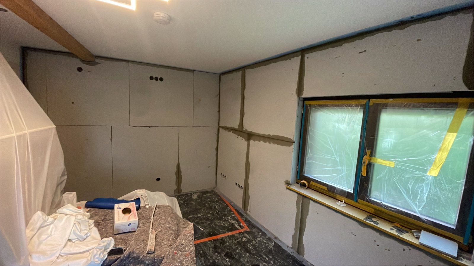 Helle Platten an Zimmerwand mit grauer Masse verklebt, Fenster abgeklebt.