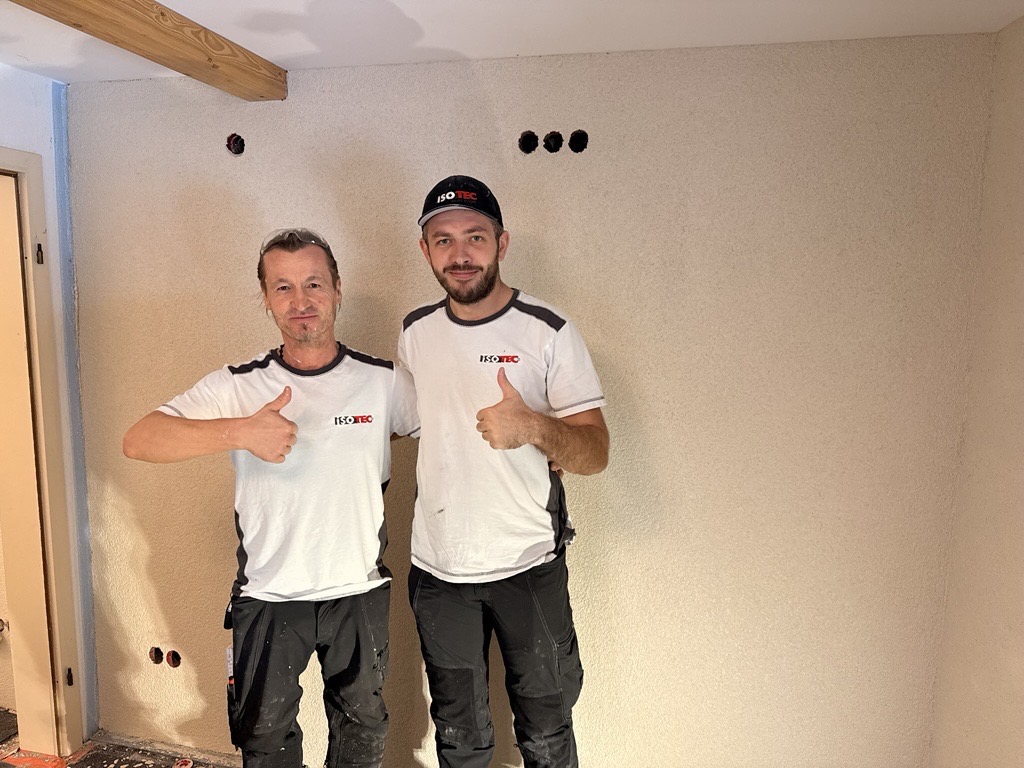 Zwei ISOTEC Mitarbeiter zeigen Daumen hoch vor heller Wand mit grober Struktur