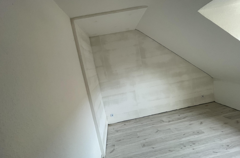 Leerer Raum mit Dachschräge, hellen Wänden und grauem Laminatboden