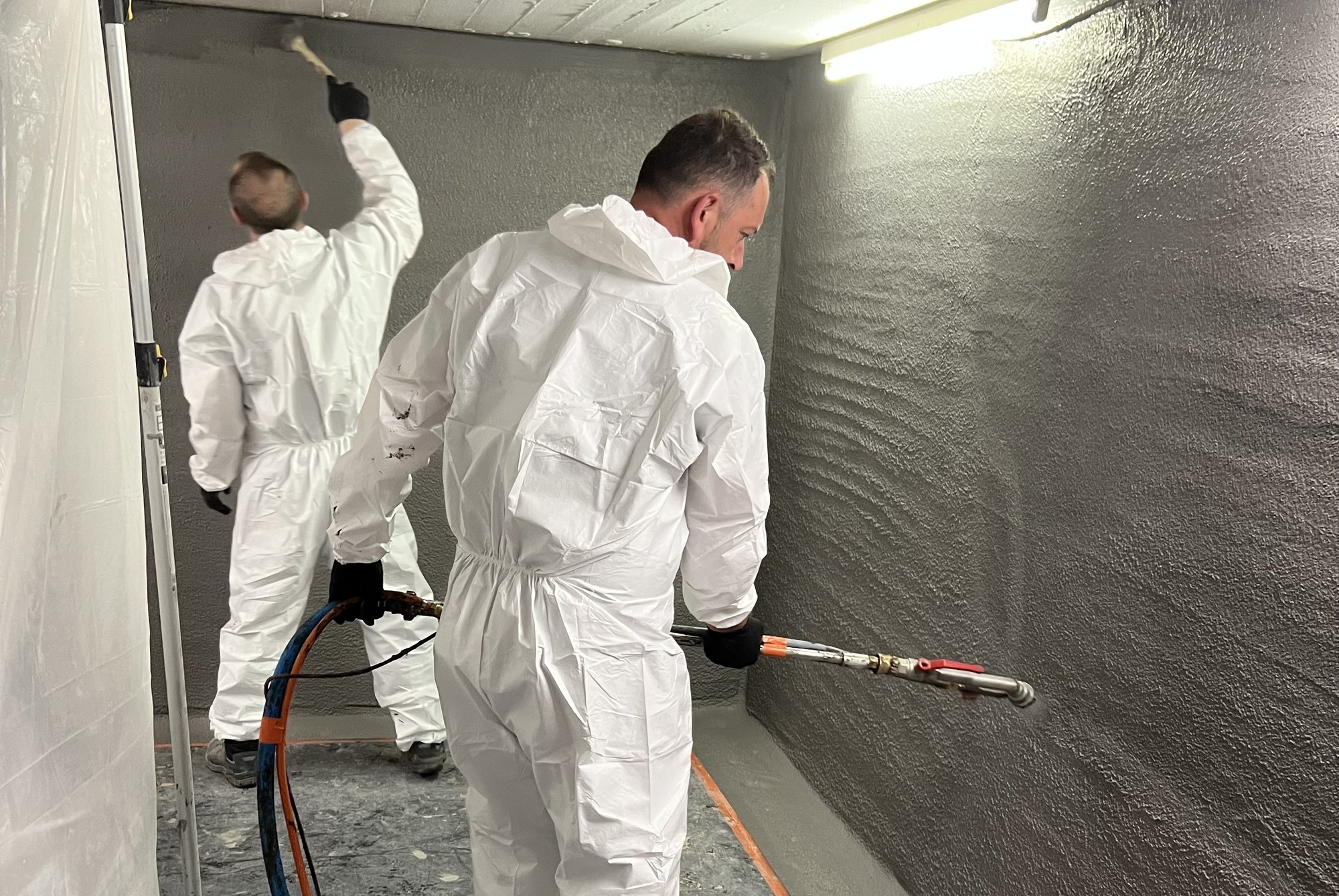 Zwei Arbeiter tragen schwarze Masse auf Kellerwände auf.