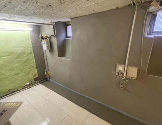 Saniertes Kellerzimmer mit grauer Beschichtung an Wand, Fliesenboden und kleinem Fenster.