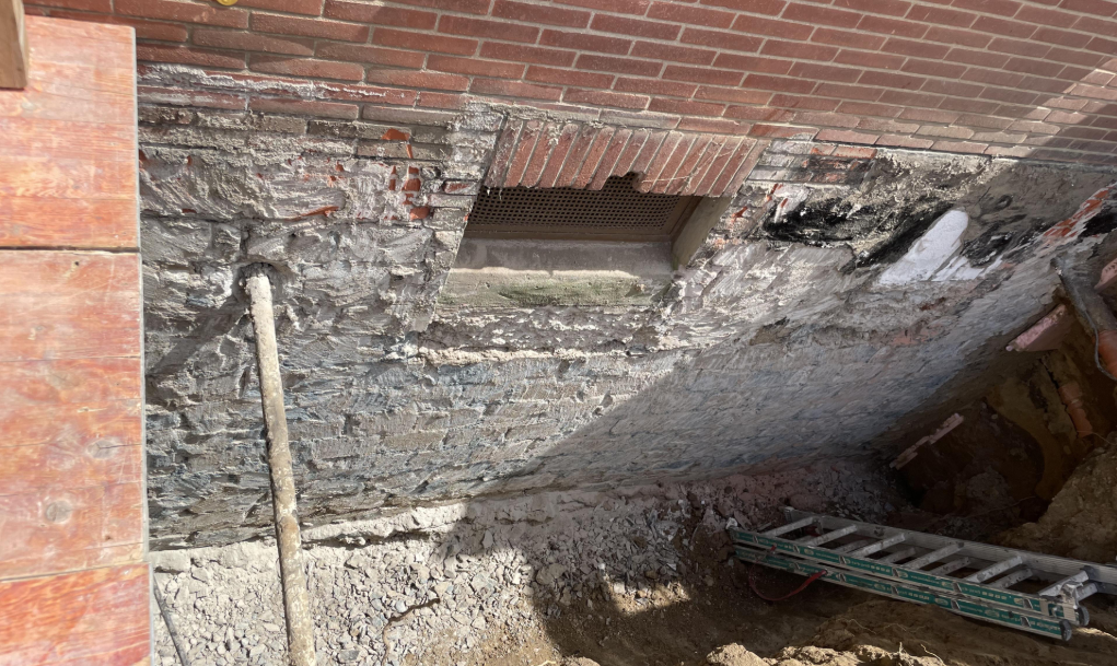 Freigelegte Ziegelwand mit Kellerfenster, grauer Masse und Bauschutt in Baugrube