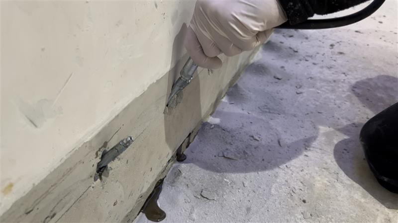 Hand mit Injektionsgerät an Kellerwand, graue Masse tritt aus Bohrlöchern aus