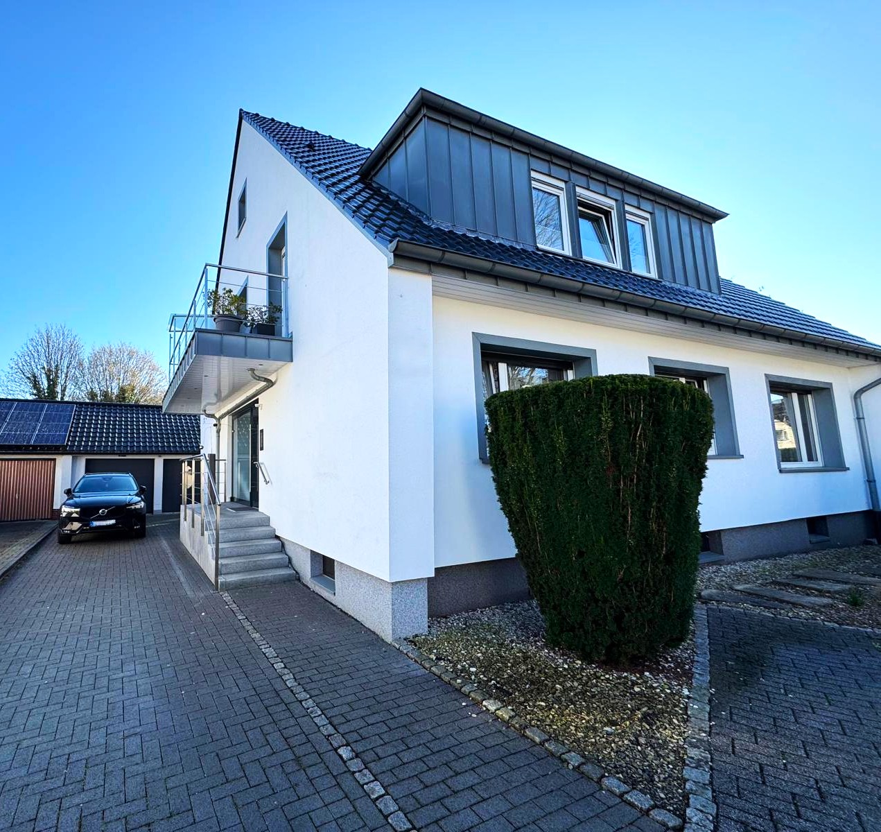 Moderne Hausfassade mit Balkon, dunklem Dach und gepflasterter Einfahrt bei blauem Himmel