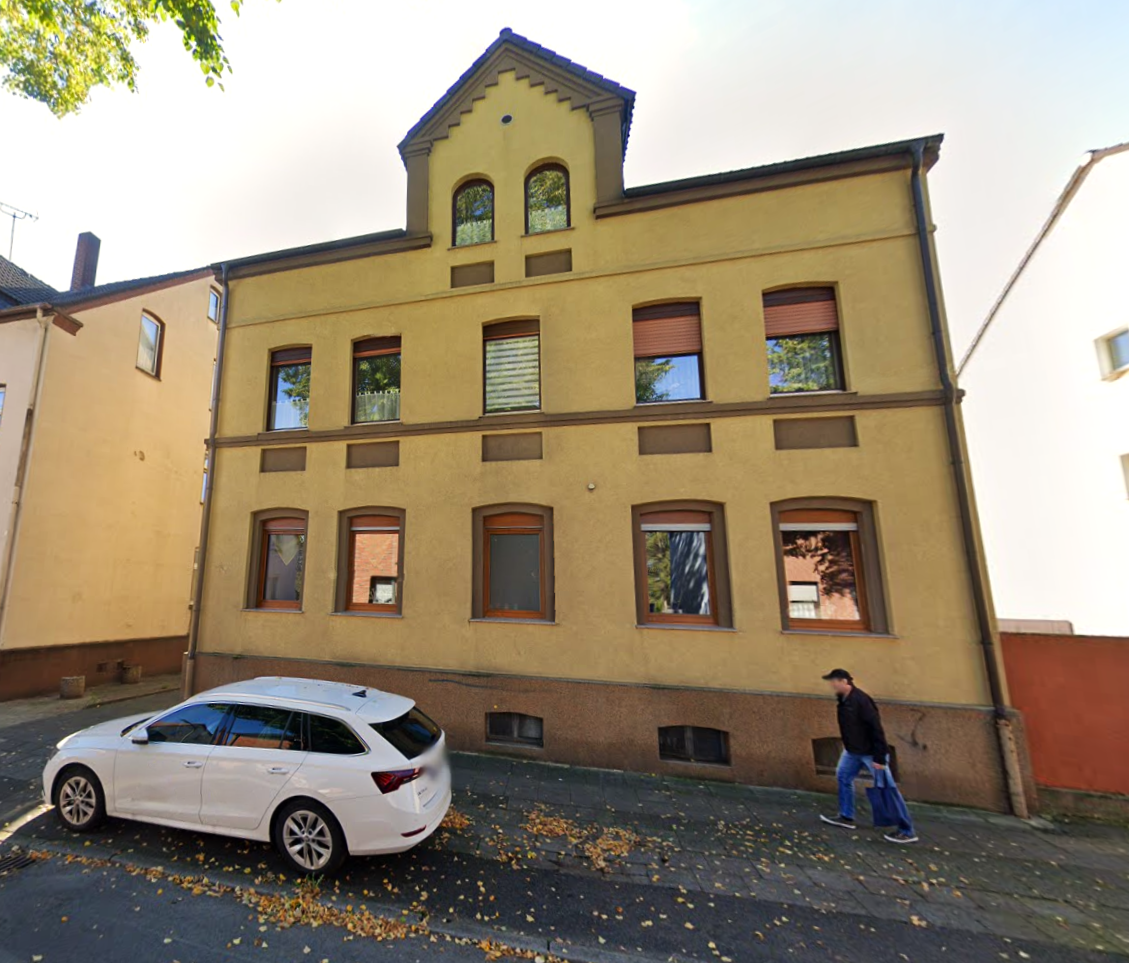 Gelbe Hausfassade mit Fenstern, parkendem Kombi und Fußgänger auf Gehweg.