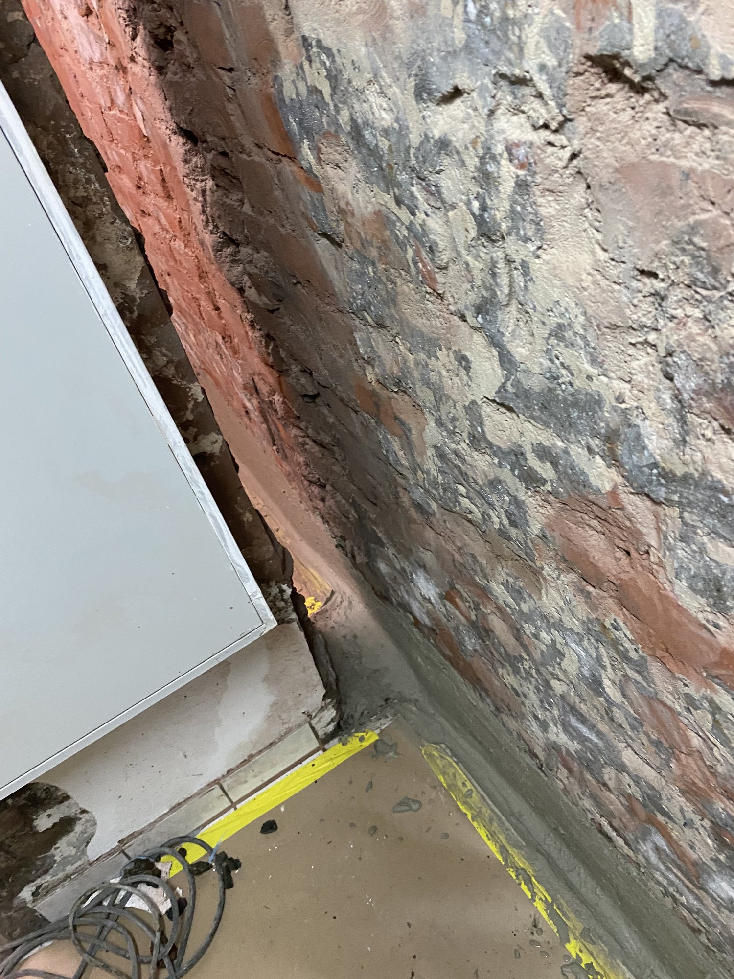 Freigelegte Kellerwand aus Mauerwerk mit grauer Masse am Wandfuß und gelbem Klebeband am Boden