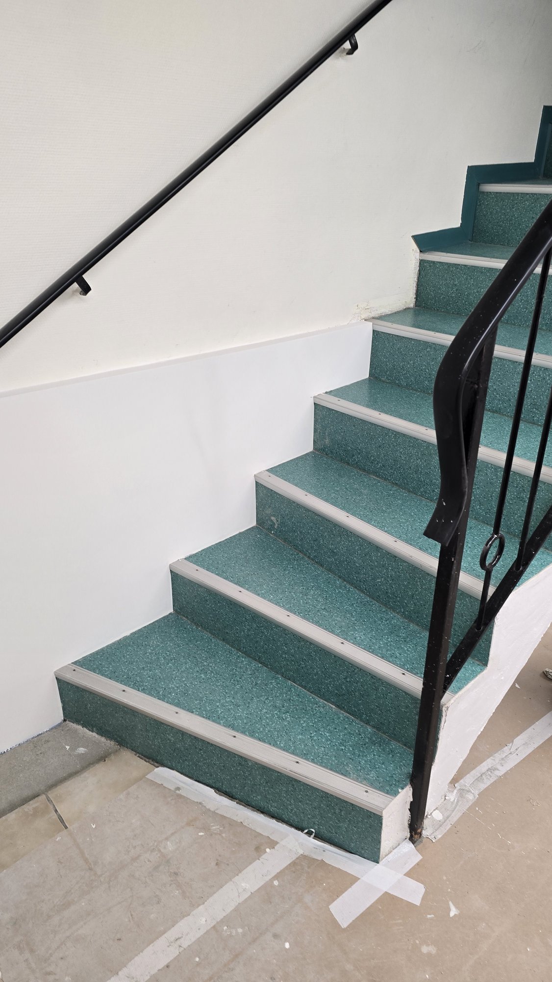 Treppenhaus mit grün belegten Stufen, schwarzem Geländer und weißer Wand