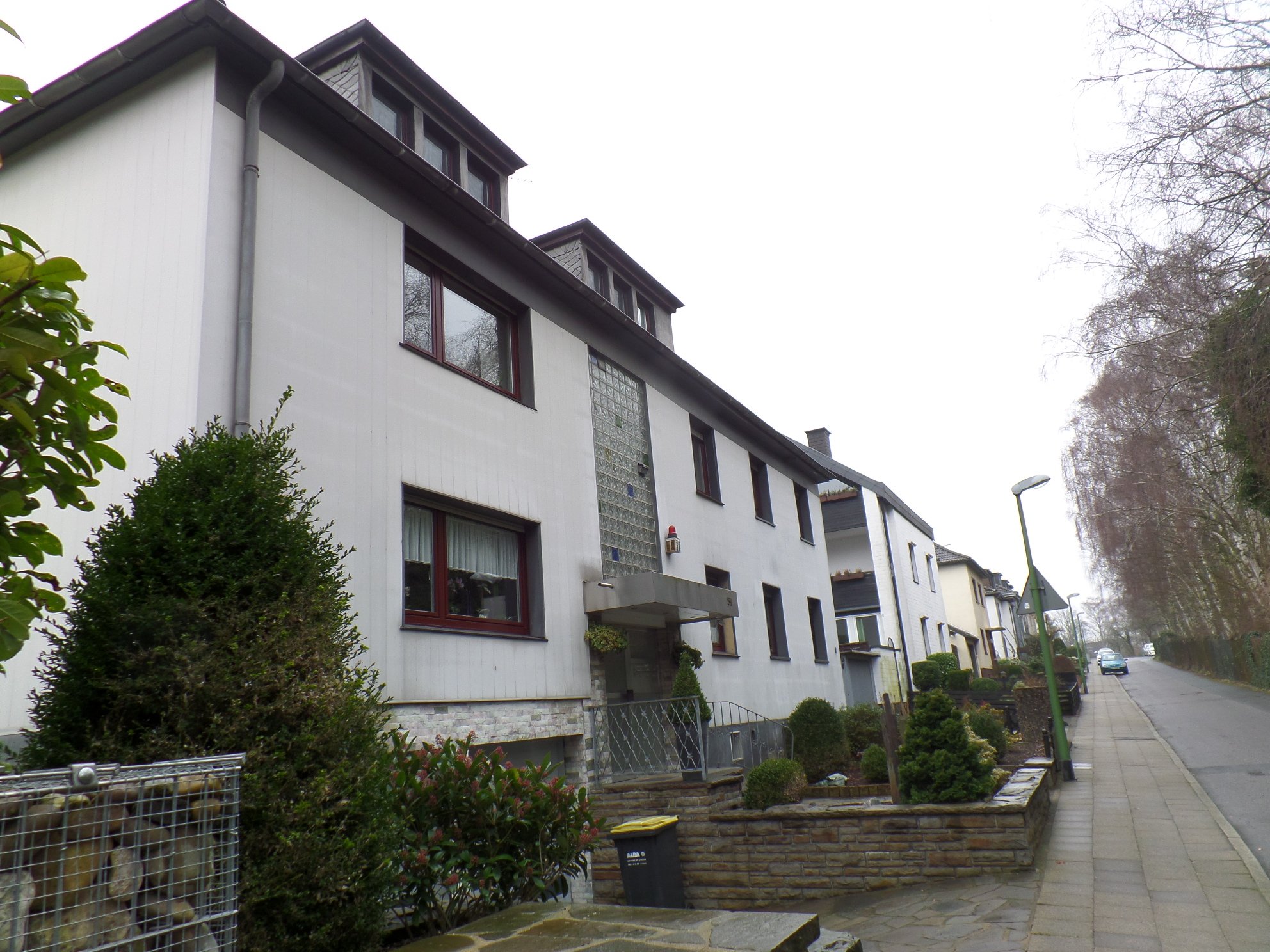 Mehrfamilienhaus mit weißer Fassade an Gehweg, Vorgarten und Straßenlaterne.