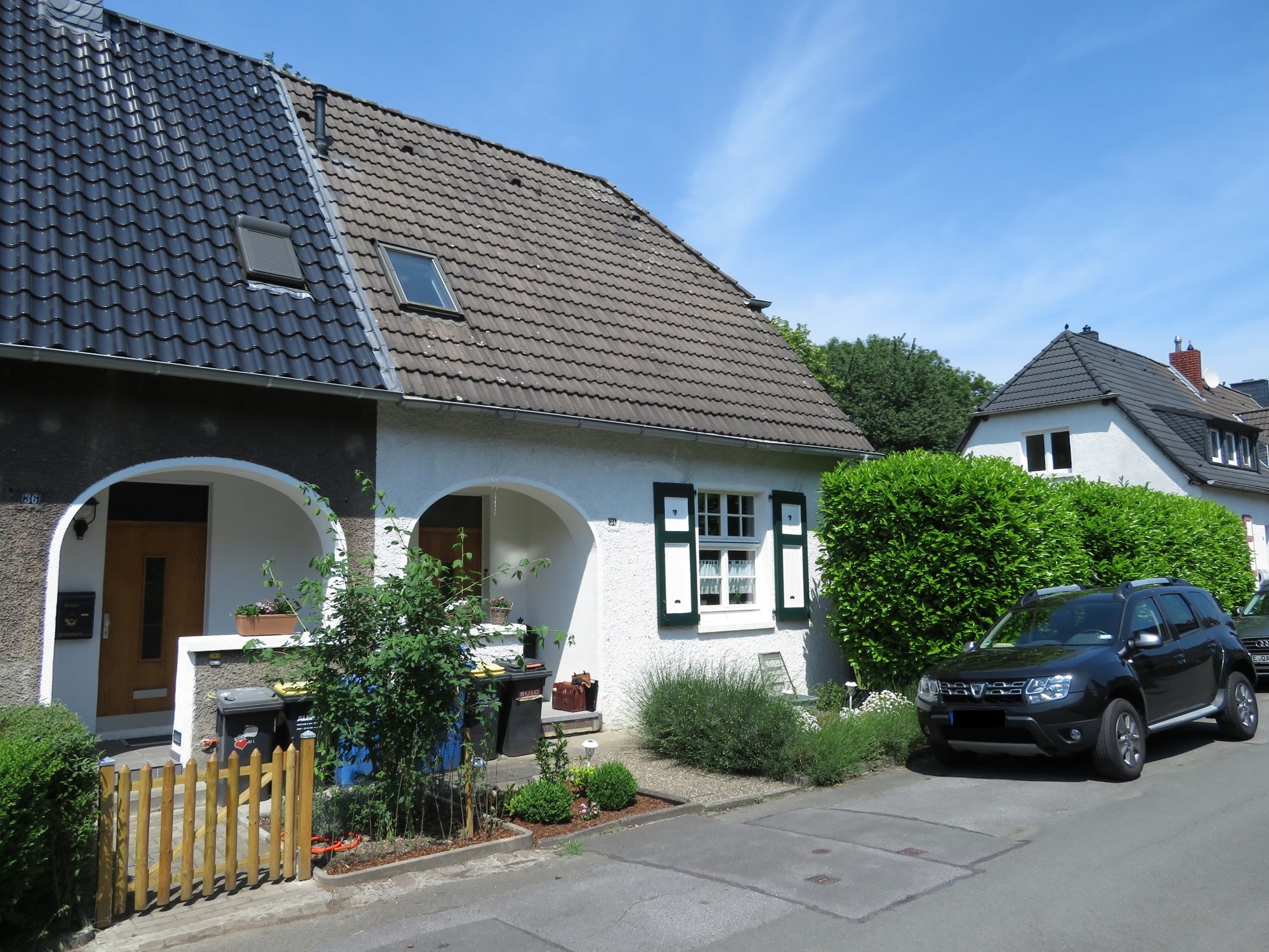Weißes Wohnhaus mit Ziegeldach, grünen Fensterläden, Hecke und geparktem Auto.