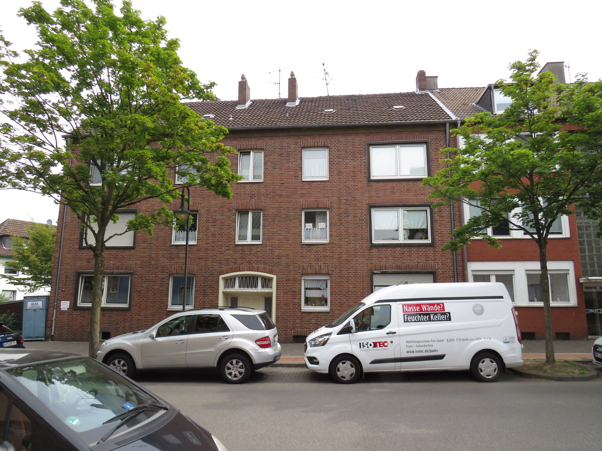 Mehrfamilienhaus aus Backstein an Straße, ISOTEC Transporter und Autos davor