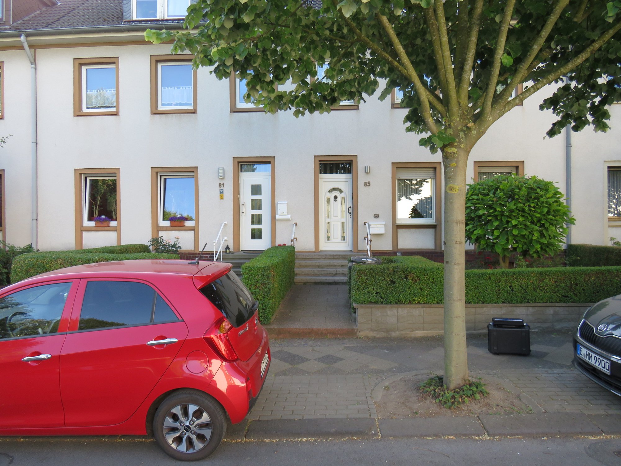 Reihenhausfassade mit zwei Haustüren, Hecken, Baum und parkendem roten Auto.