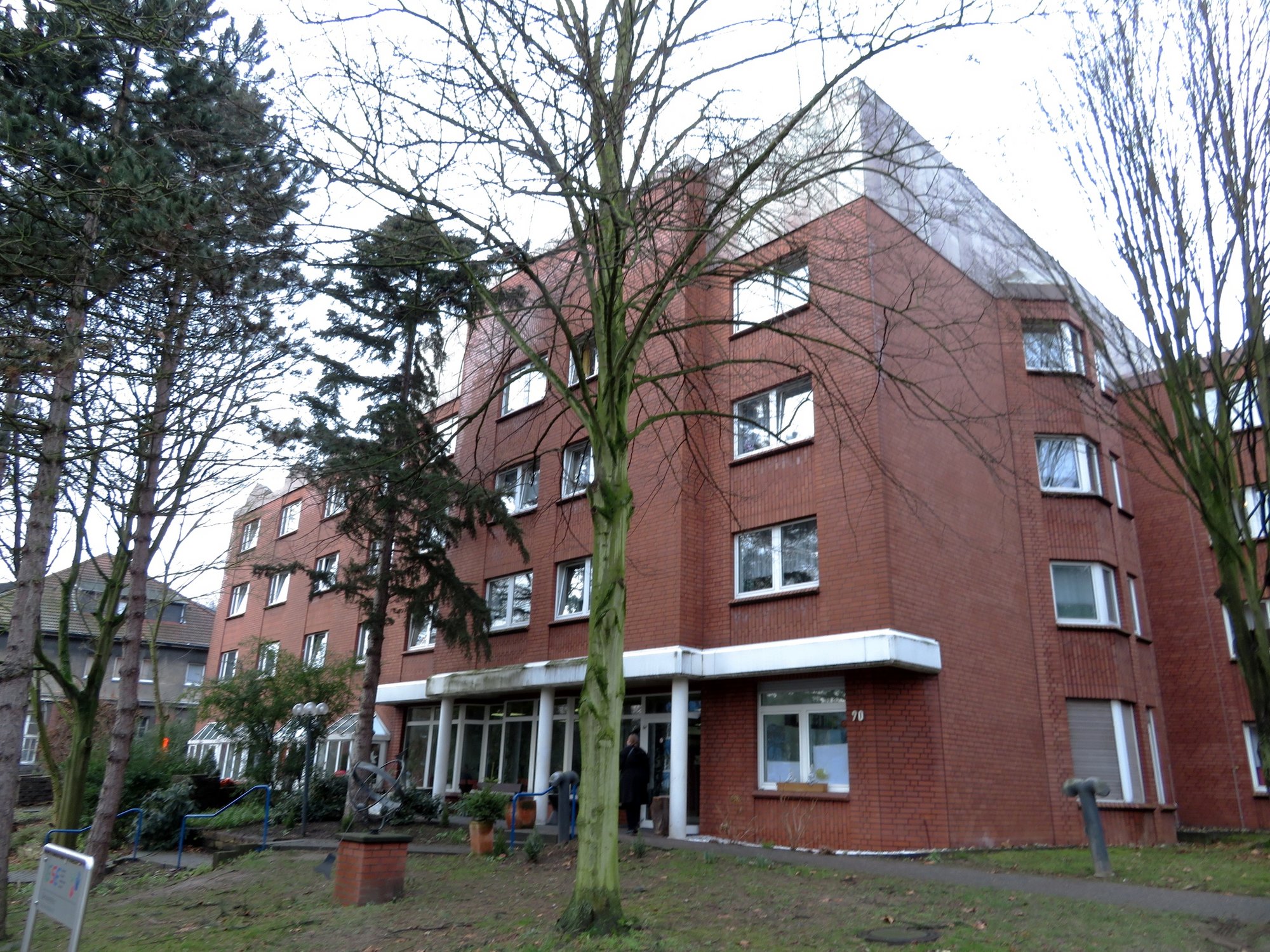 Mehrstöckiges Backsteingebäude mit Eingangsbereich, davor kahle Bäume und Gehweg.