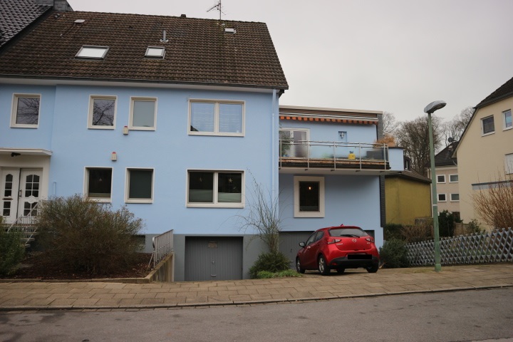Blaues Mehrfamilienhaus mit Balkon, Garageneinfahrt und rotem Auto an der Straße
