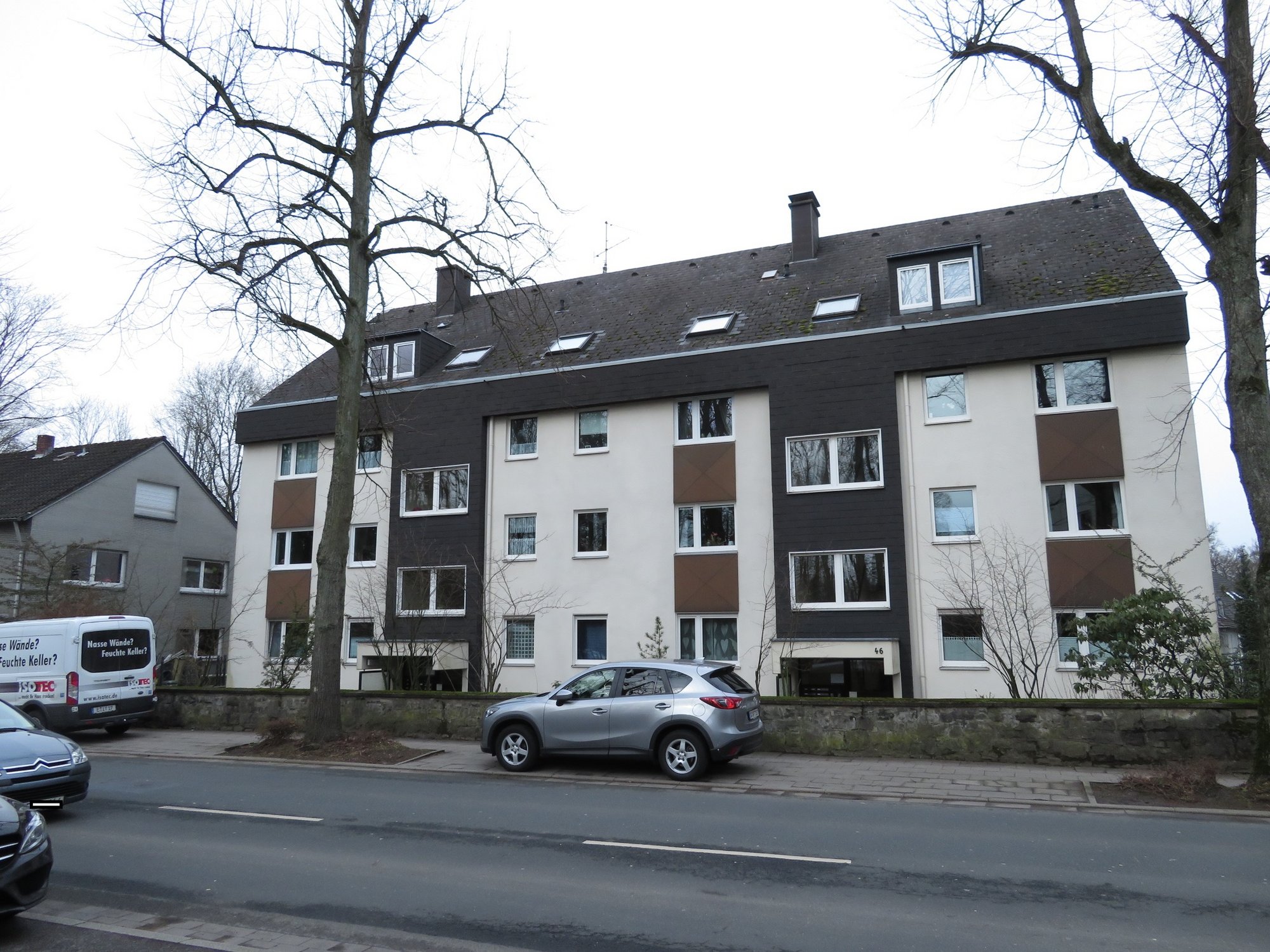 Mehrfamilienhaus an Straße mit geparkten Autos und kahlen Bäumen, Frontansicht