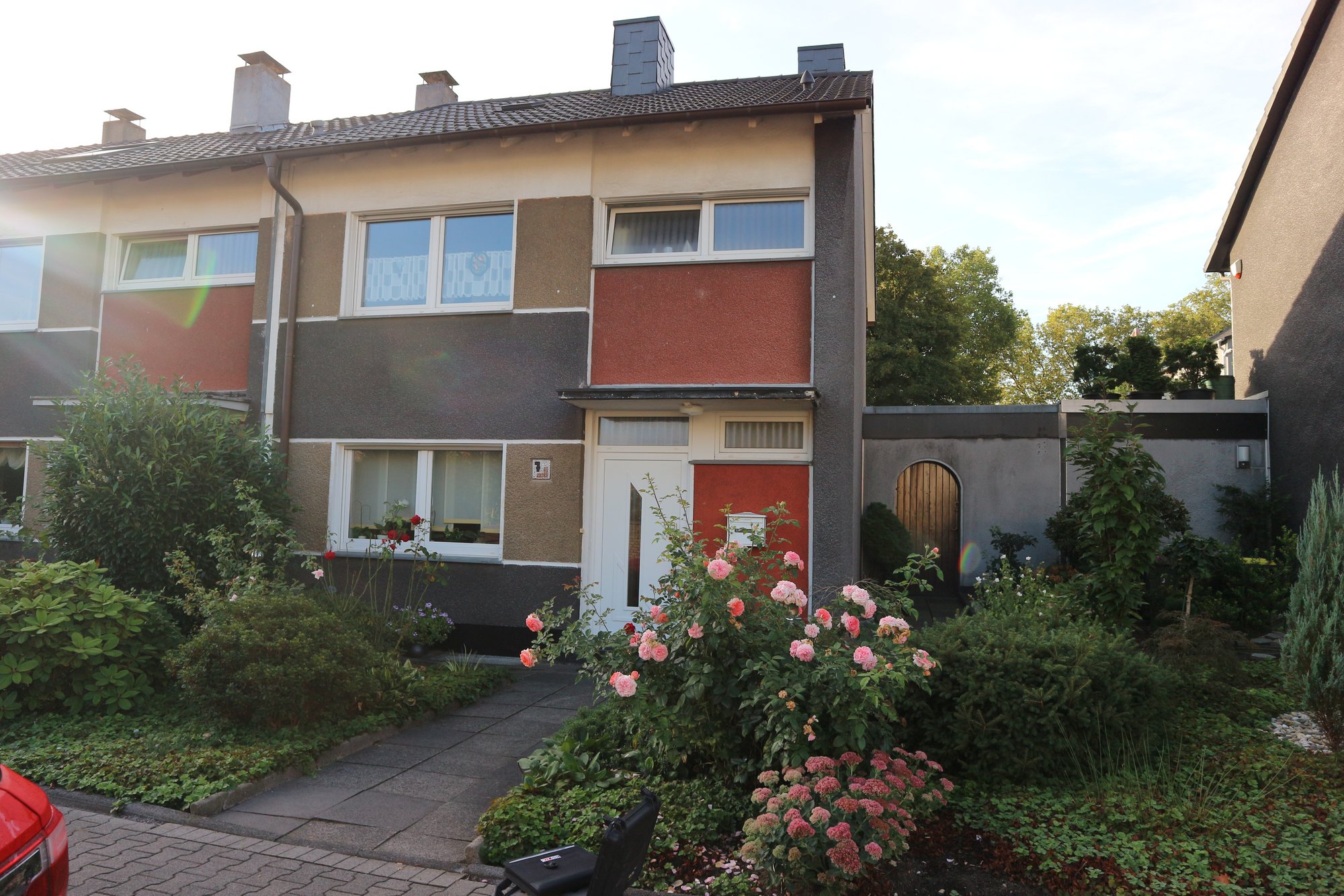 Reihenhausfassade mit weißen Fenstern und Eingangstür, davor Garten mit Rosen und Gehweg.