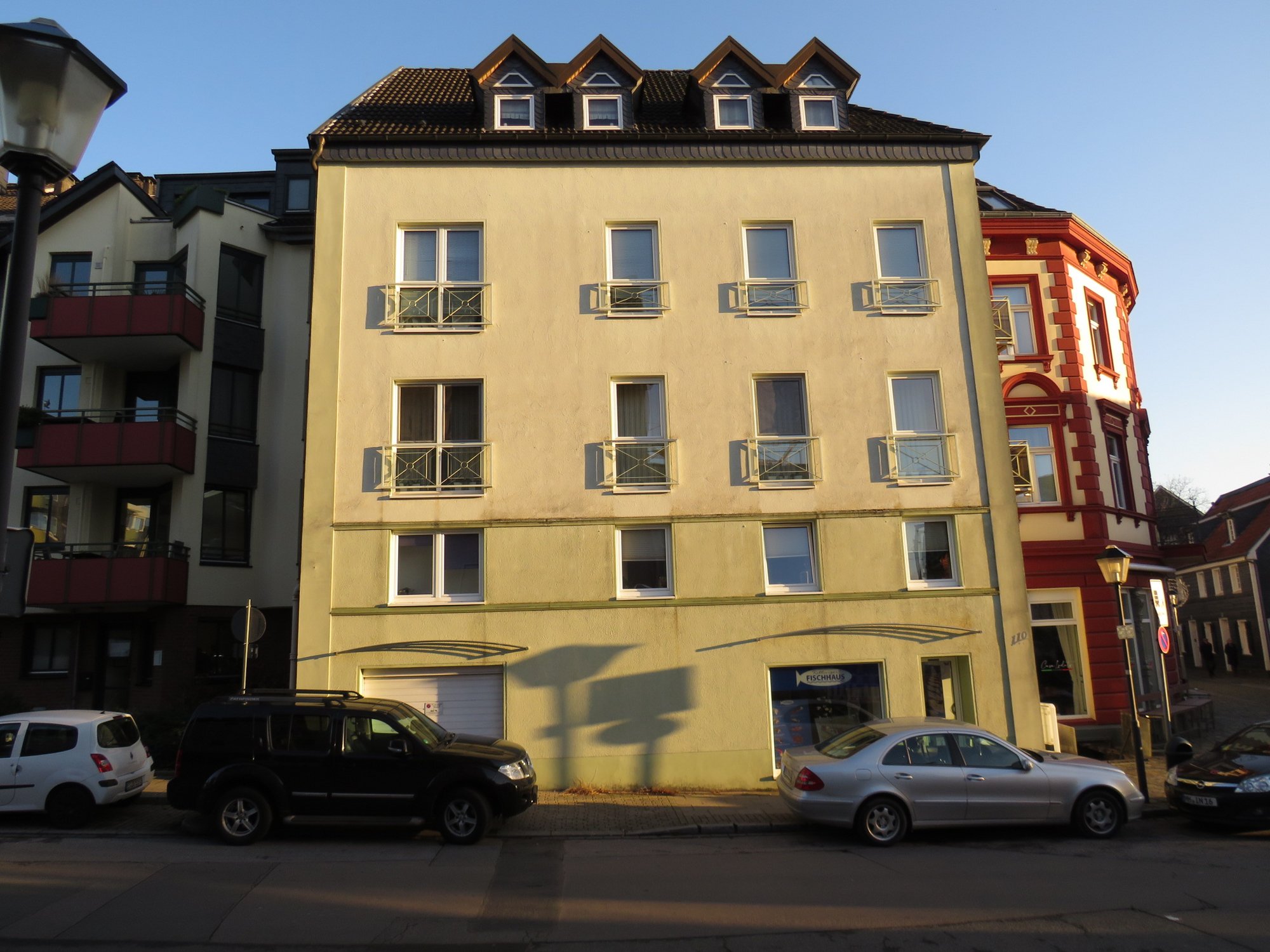 Mehrstöckiges Wohnhaus mit Balkonen und parkenden Autos in Straßenansicht.