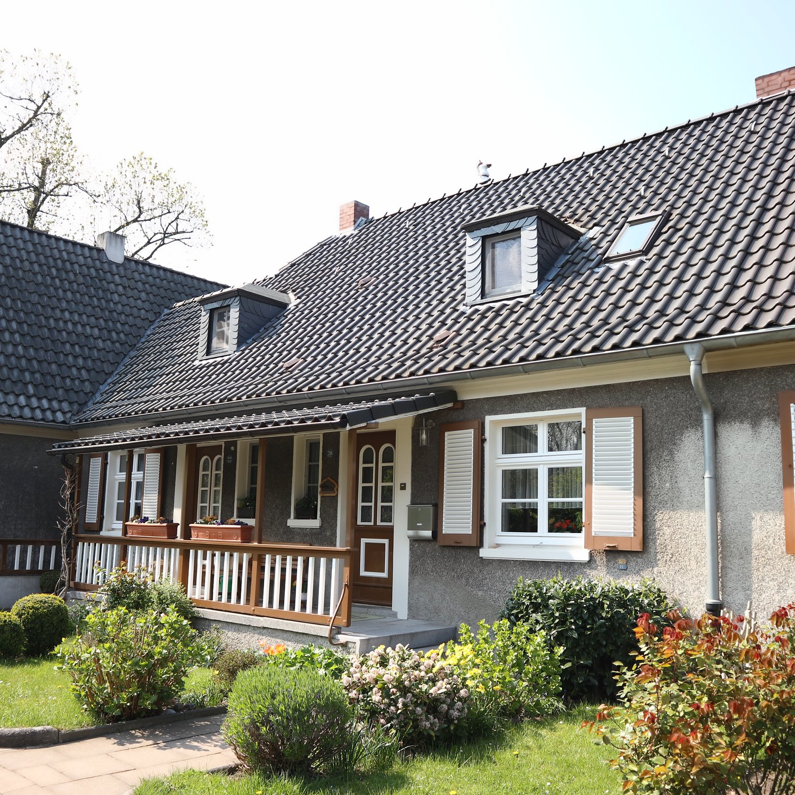Einfamilienhaus mit dunklem Ziegeldach, Gaubenfenstern, Fensterläden und Vorgarten.