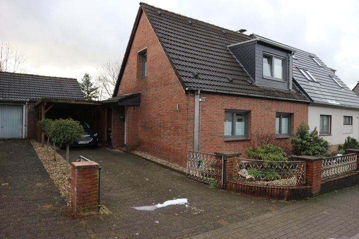 Backsteinhaus mit dunklem Ziegeldach, Einfahrt und Carport neben kleinem Vorgarten