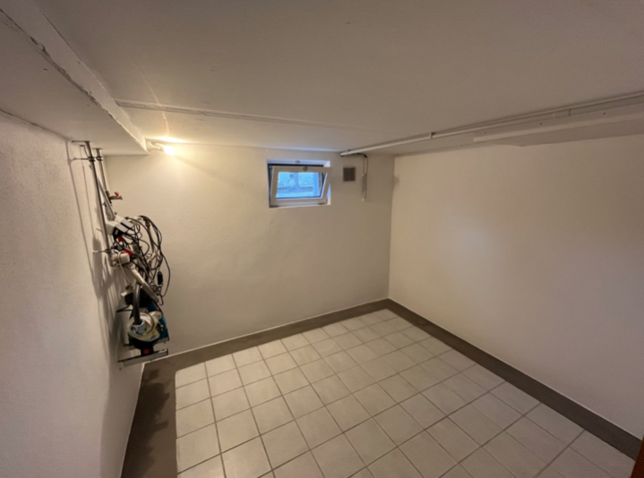 Leerer Kellerraum mit Fliesenboden, kleinem Fenster und freiliegender Haustechnik