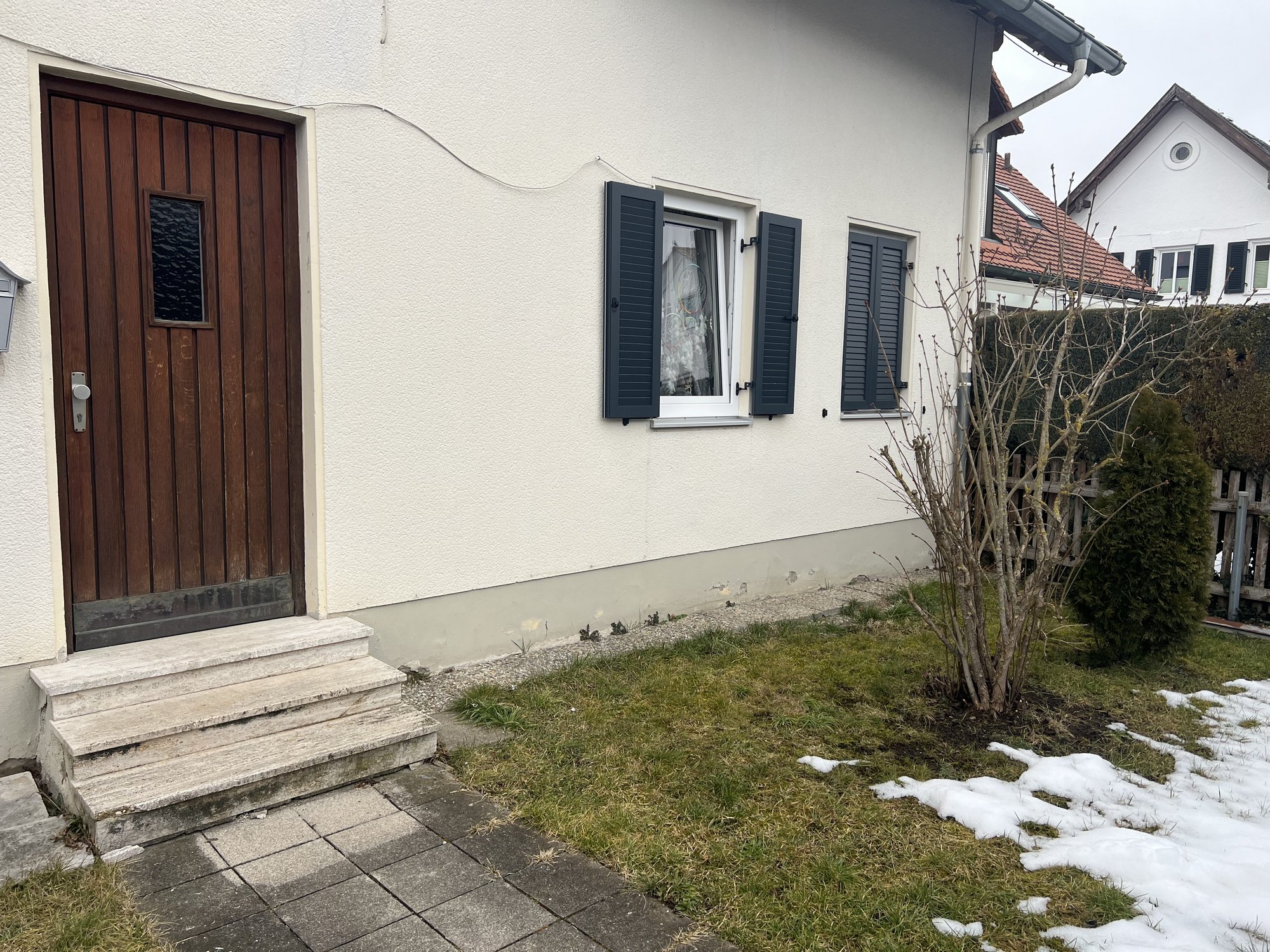 Hauseingang mit Holzhaustür, weißen Fassadenfenstern und Garten mit Restschnee
