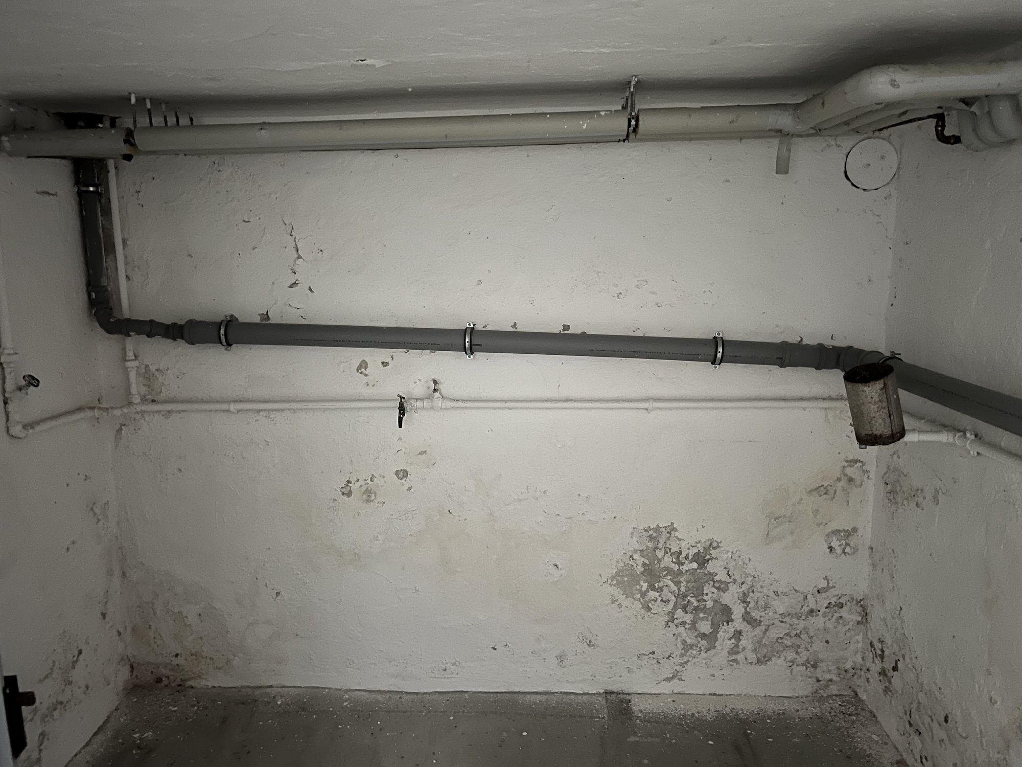 Abplatzender Anstrich an Kellerwand mit grauen Rohrleitungen und Ventil