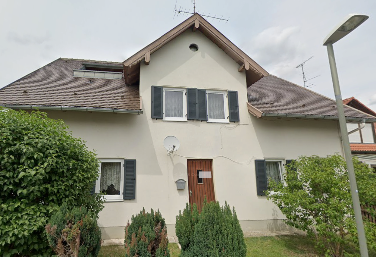 Vorderansicht eines Hauses mit Satteldach, Fensterläden, Holzhaustür und Sträuchern