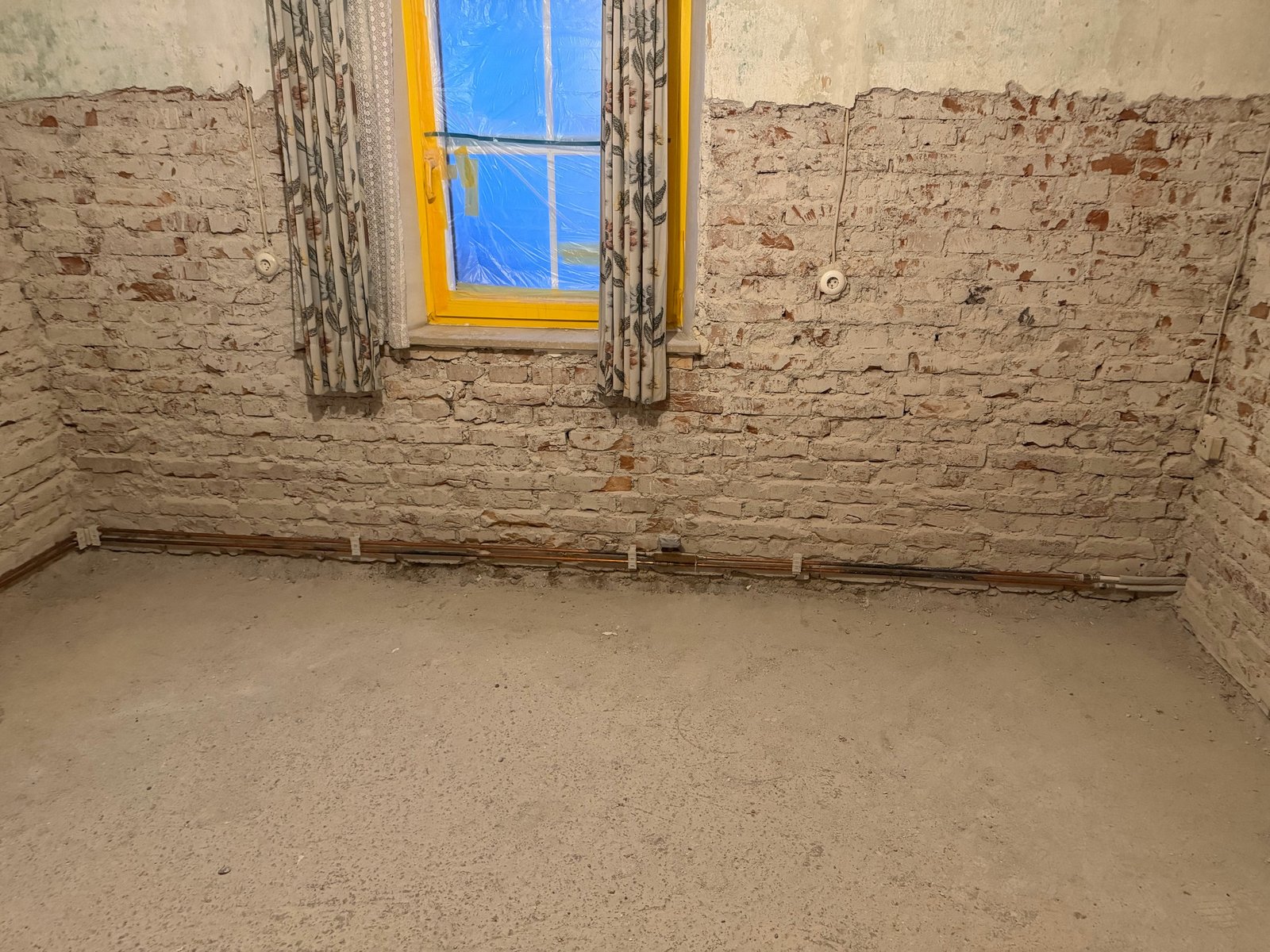 Freigelegte Ziegelwand in leerem Raum mit Fenster und sichtbaren Leitungen