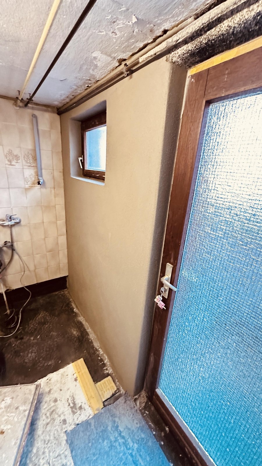 Tür und kleines Fenster in gefliestem Raum, Decke mit Abplatzungen und Rohrleitungen.