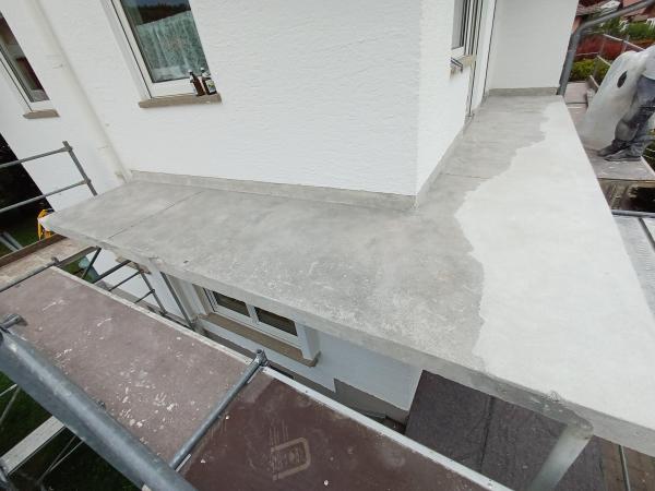 Grauer Balkonboden mit abgegrenzter, dunklerer Fläche neben weißer Fassade.