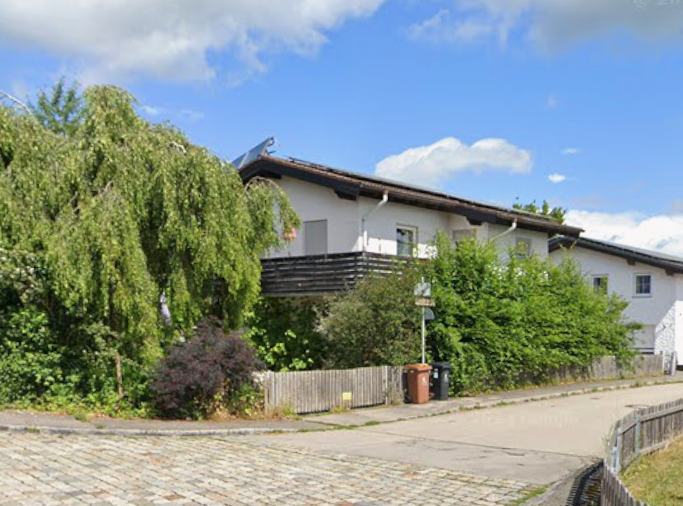 Wohnhaus mit Balkon, Bäumen und Holzzaun an ruhiger Straße unter blauem Himmel