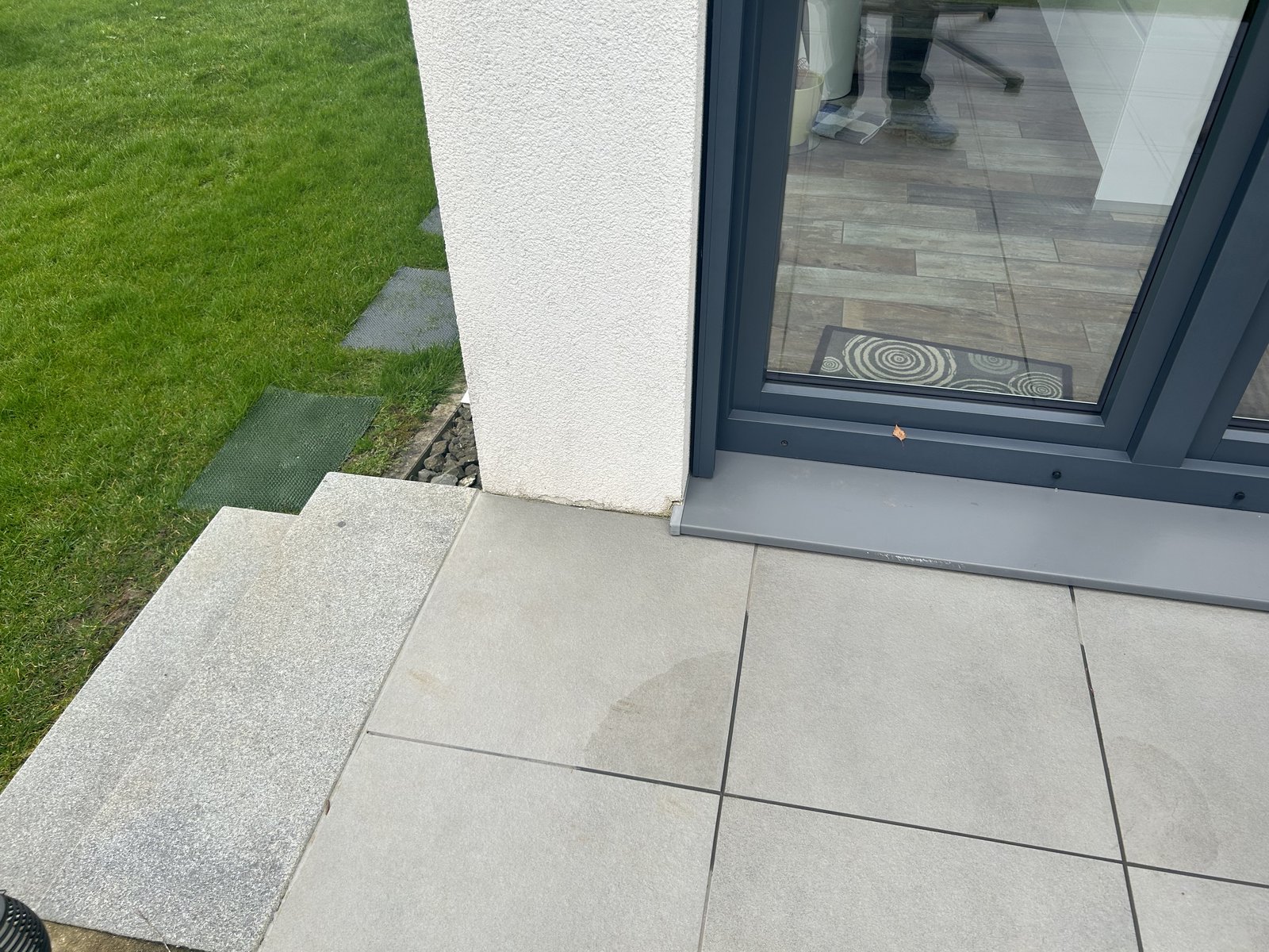 Terrassenfliesen vor bodentiefem Fenster, daneben Rasen und Steinstufe.