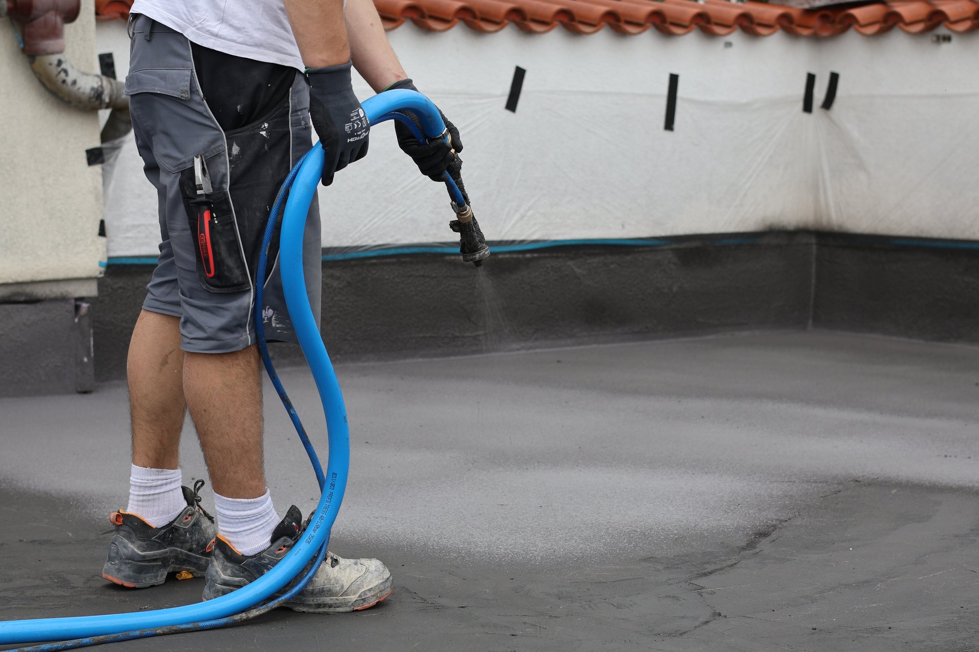 Arbeiter sprüht transparente Flüssigkeit auf graue Dachfläche mit Schlauchdüse.