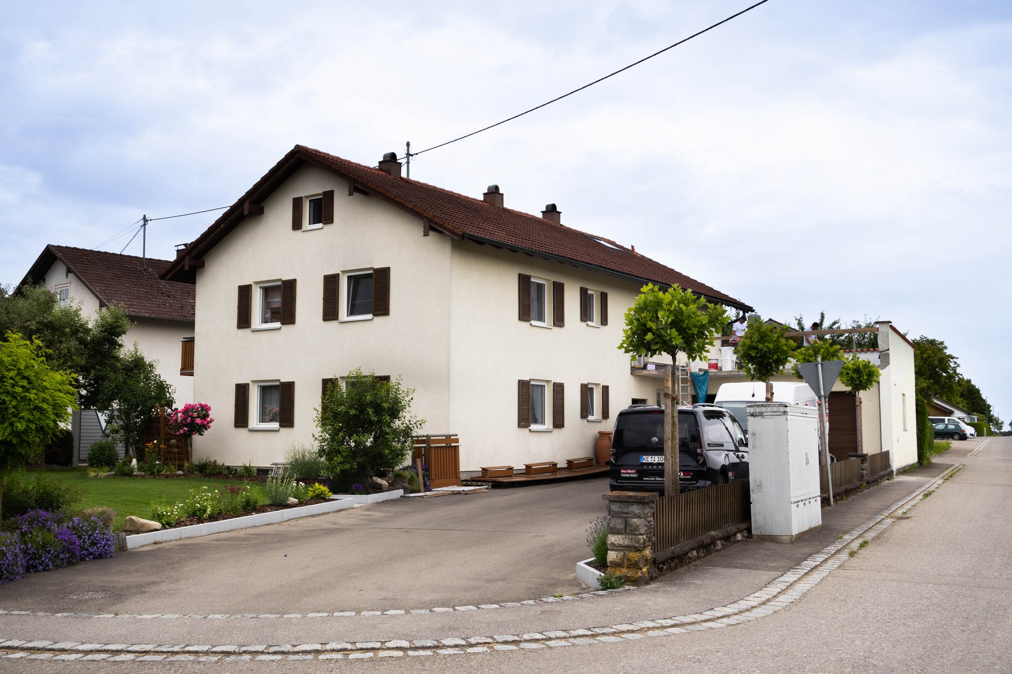 Wohnhaus mit rotem Ziegeldach, Garten und geparkten Fahrzeugen an einer Straße