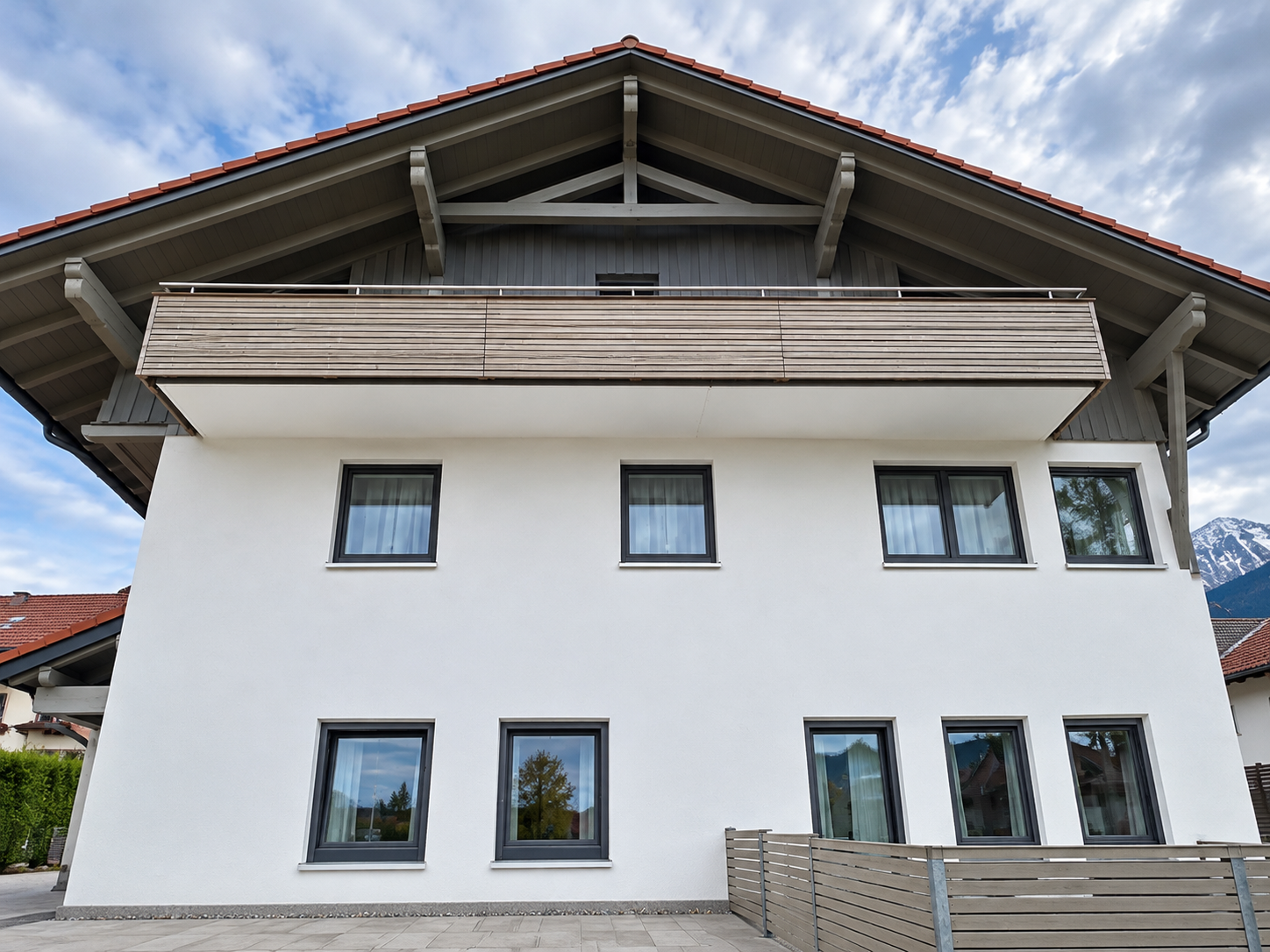 Gebäude mit weißer Fassade, Holzbalkon und dunkelgrauen Fenstern unter rotem Ziegeldach