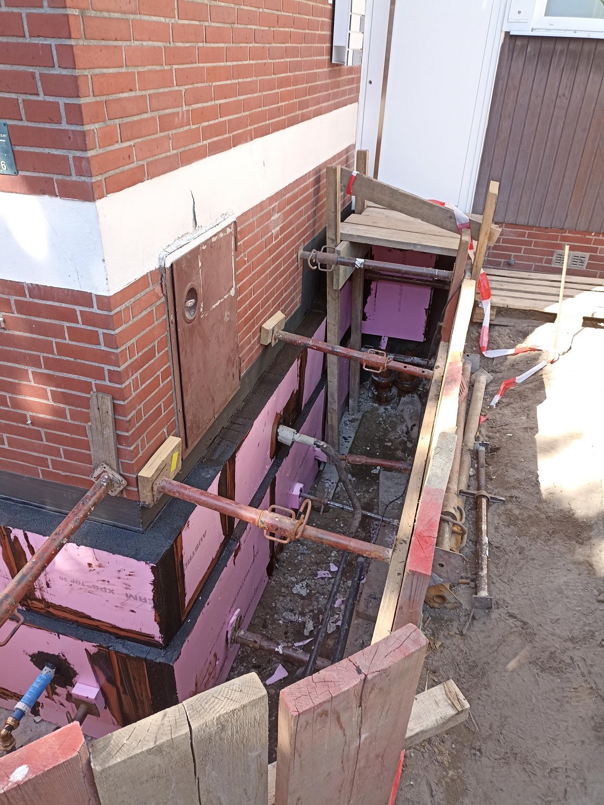 Baugrube mit Schalung und rosa Dämmplatten an Gebäudesockel aus Klinker