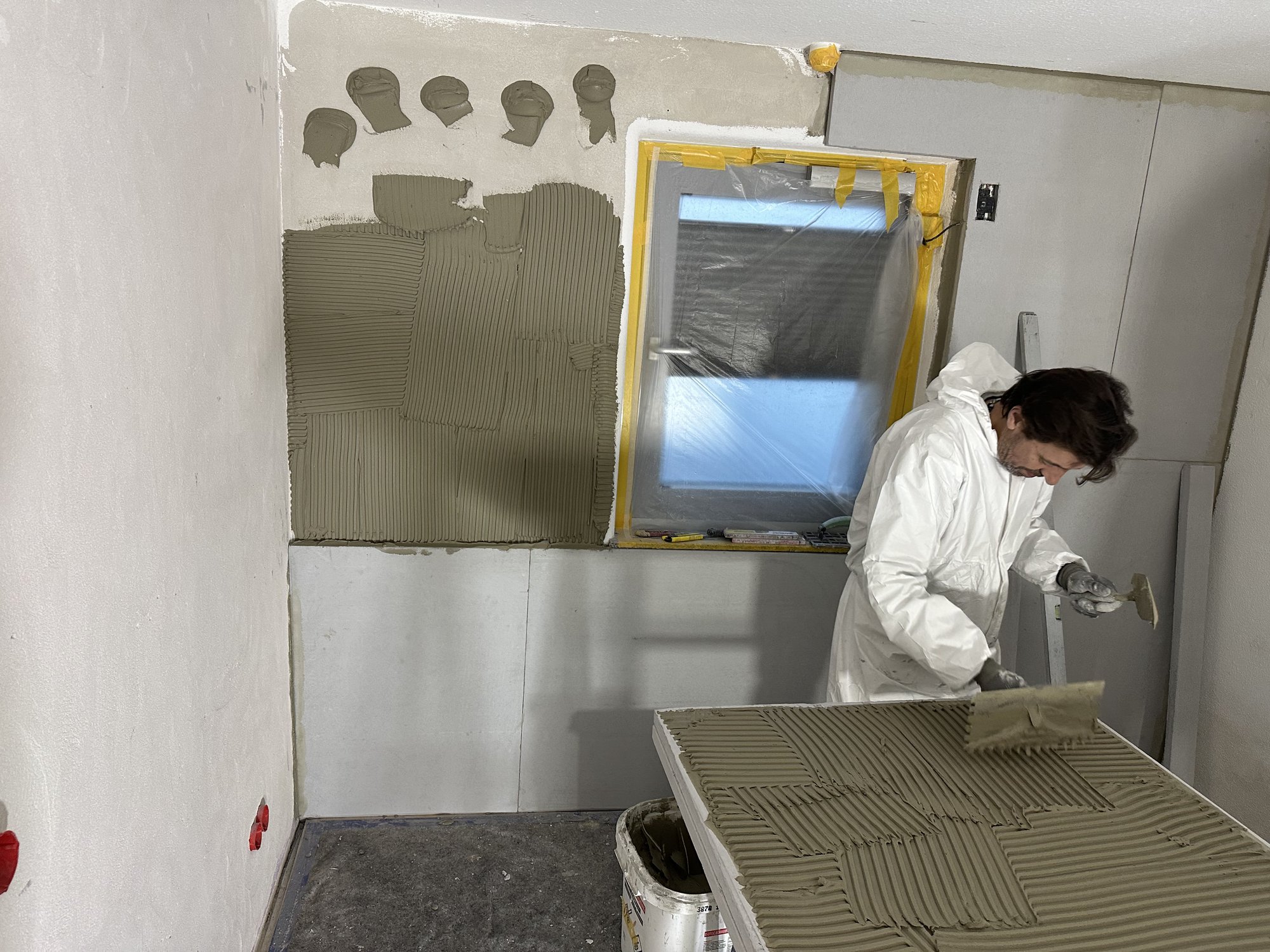 Arbeiter trägt graue Masse mit Zahnspachtel auf helle Platte auf, abgeklebt­es Fenster daneben.