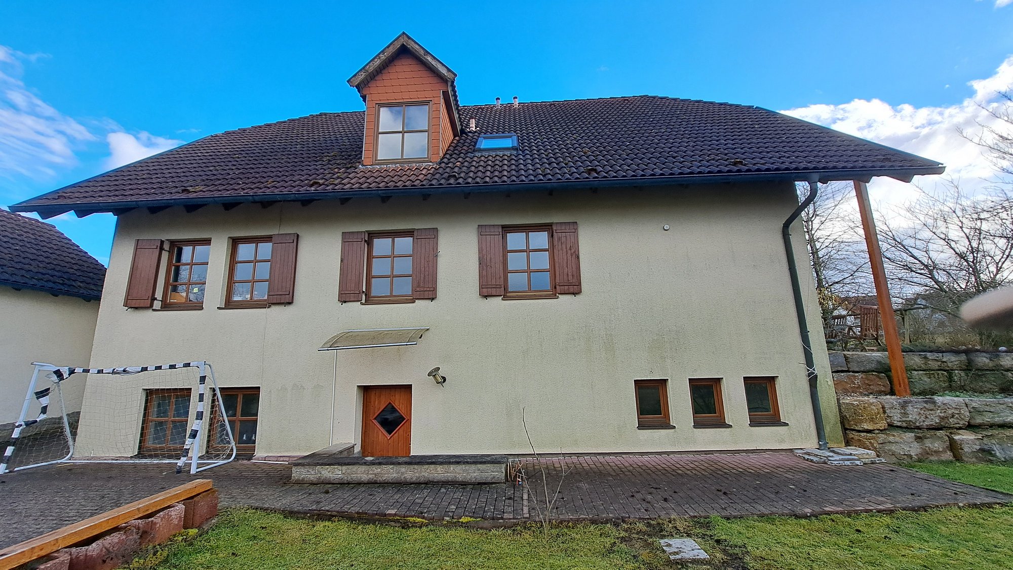 Zweigeschossiges Haus mit braunem Ziegeldach, Holzfenstern und gepflasterter Terrasse