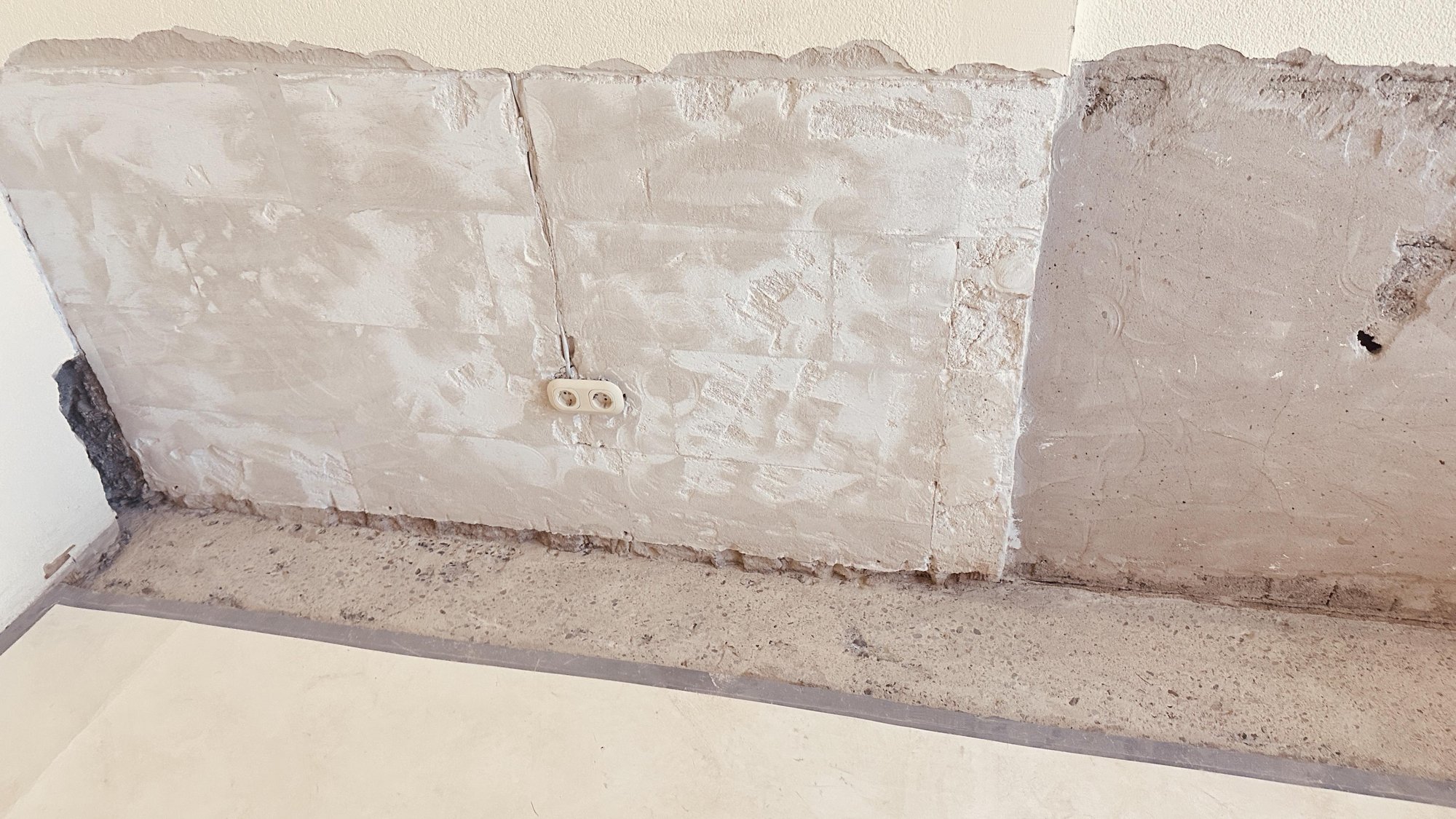 Freigelegte Betonwand mit grauer Masse und Steckdose, Sockelbereich beschädigt