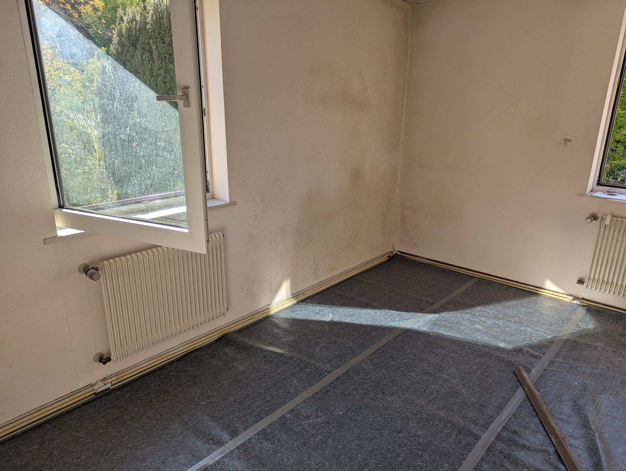 Fleckige Zimmerecke mit dunklen Wandverfärbungen, Fenster und Heizkörper, Boden mit Schutzvlies.