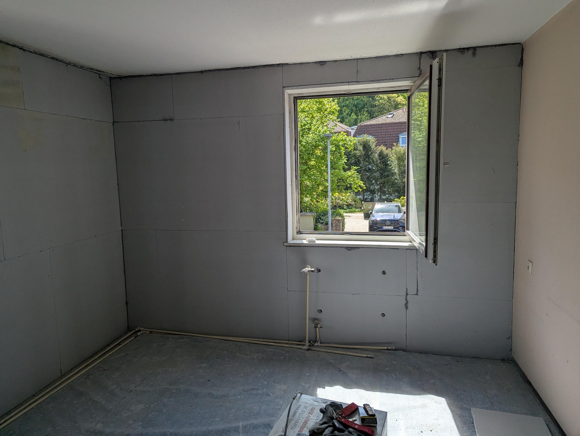 Unfertiger Raum mit grauen Plattenwänden, offenem Fenster und sichtbaren Rohren am Boden