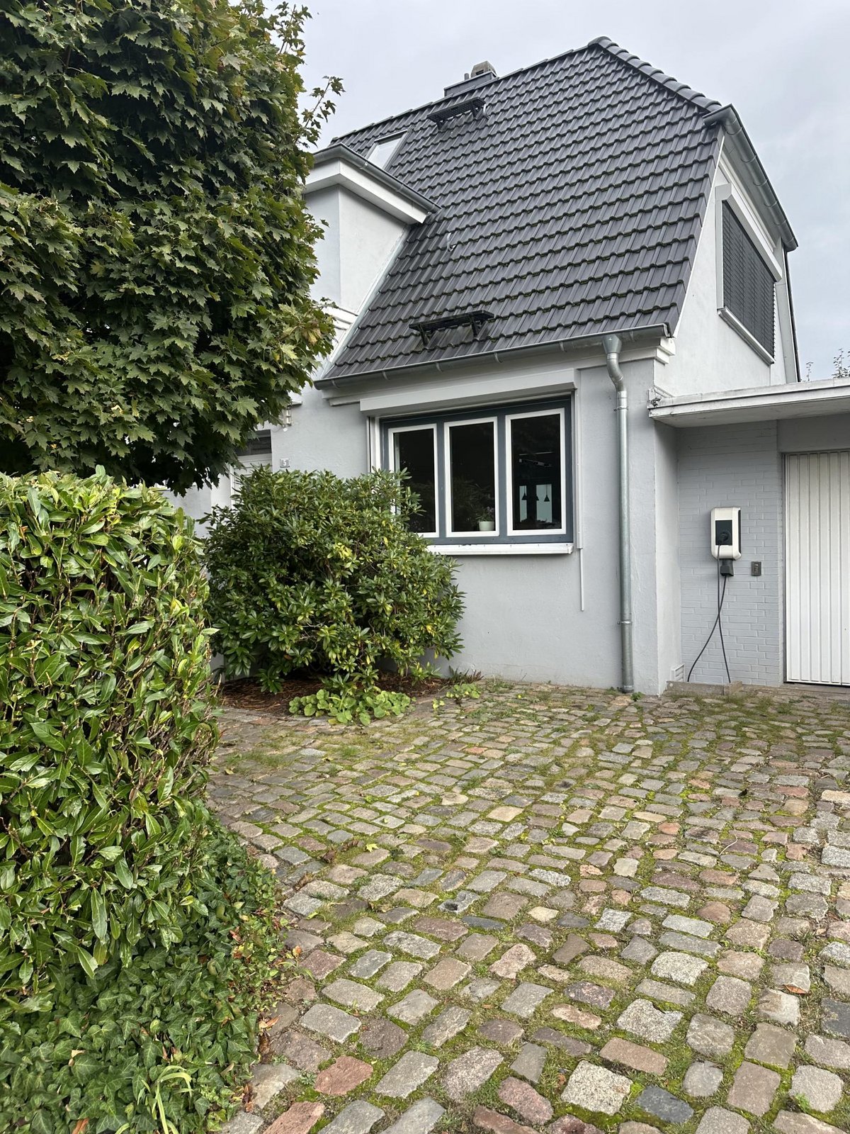 Hausfassade mit dunklem Ziegeldach, Einfahrt aus Kopfsteinpflaster und Sträuchern