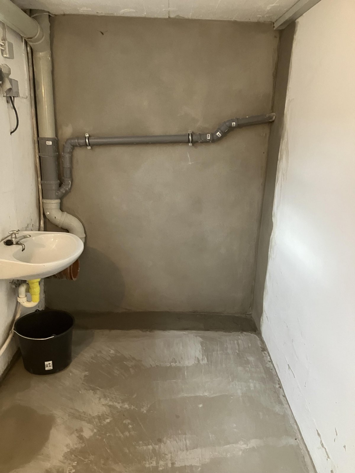Kellerraum mit grauer Wand, Abflussrohren, Waschbecken und Eimer auf Betonboden