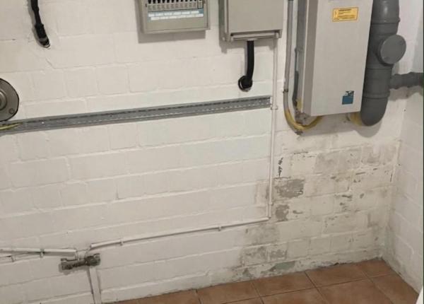 Weiße Kellerwand aus Mauerwerk mit grauen Feuchtflecken, Leitungen und Elektroverteilung, Fliesenboden
