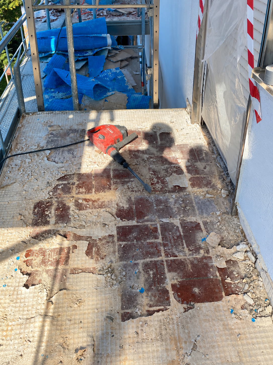 Roter Stemmhammer liegt auf staubigem Fliesenboden, Bauschutt und Gerüst am Balkonzugang