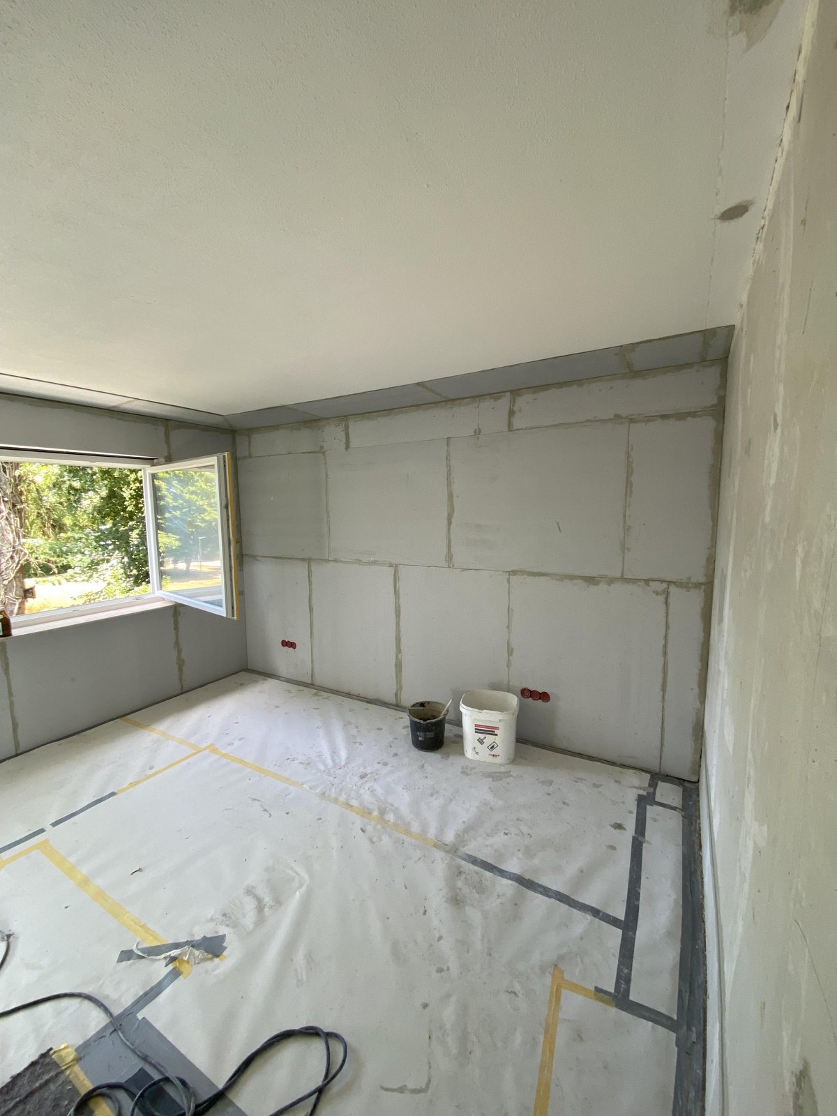 Renovierungsraum mit grauen Wandplatten, offenem Fenster und Eimern auf abgedecktem Boden