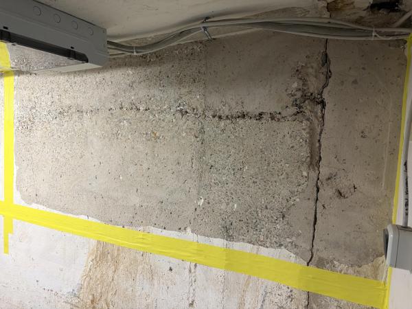 Rissige Betonwand im Keller mit dunklen Feuchtespuren und gelbem Absperrband