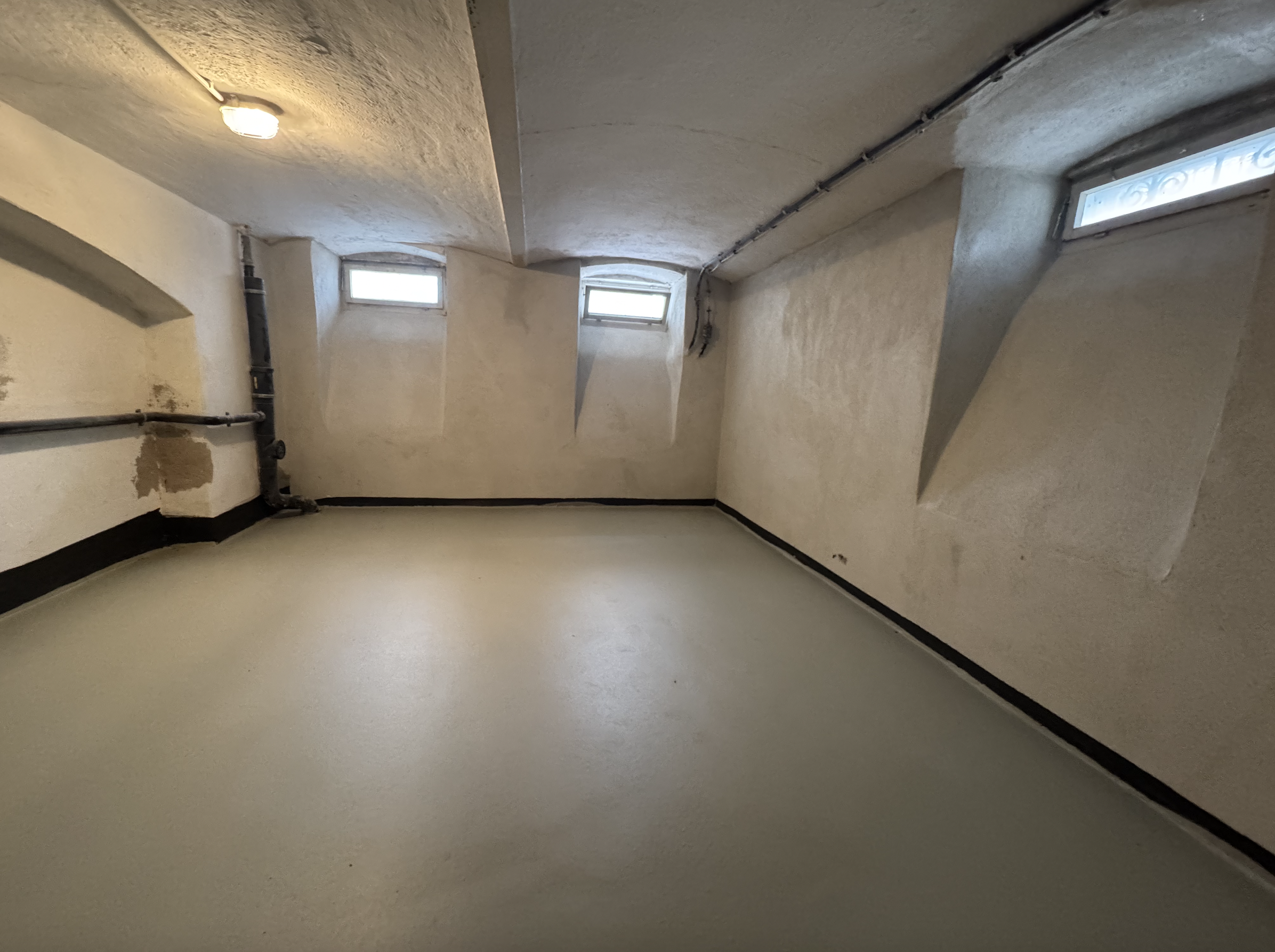 Leerer Kellerraum mit hellem Boden, kleinen Fenstern und Rohrleitung an der Wand