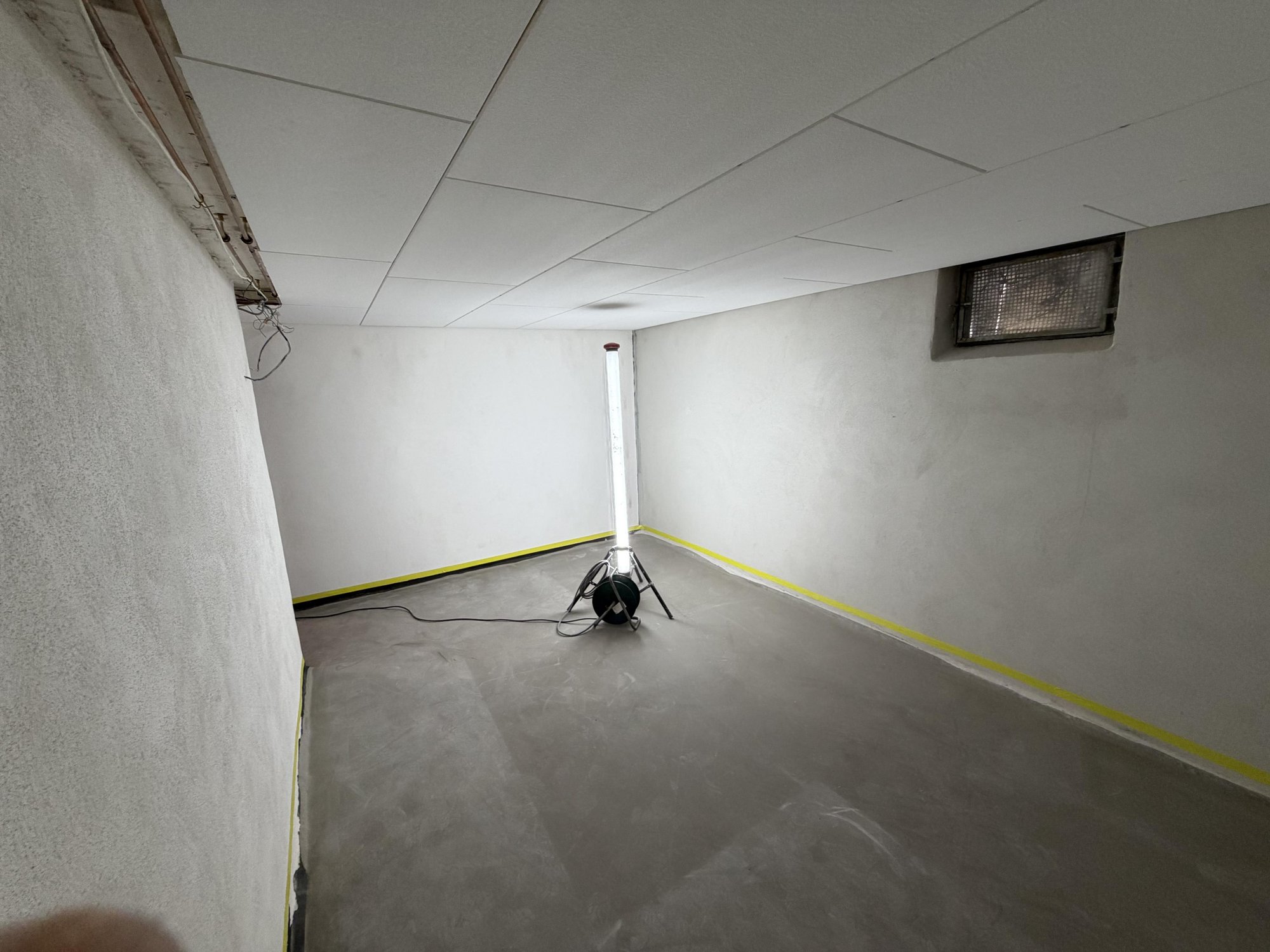 Leerer Kellerraum mit grauem Boden, weißer Decke und Baustrahler auf Stativ